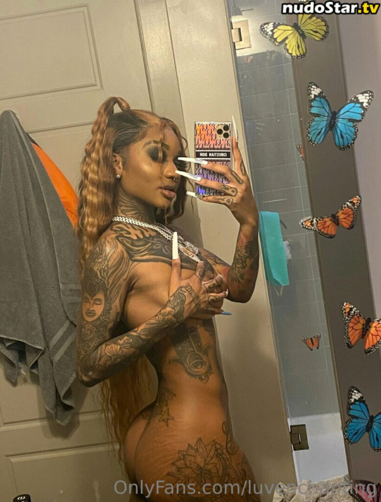 Enchanting, the OnlyFans model and rapper, takes a mirror selfie in a bathroom. She is nude, covering her chest with one hand while holding her phone with the other. Her long, wavy hair is styled in a high ponytail, and she is adorned with intricate tattoos across her body. Butterflies are visible on the bathroom mirror, adding a whimsical touch to the scene. She exudes confidence and allure, showcasing her unique style and presence.