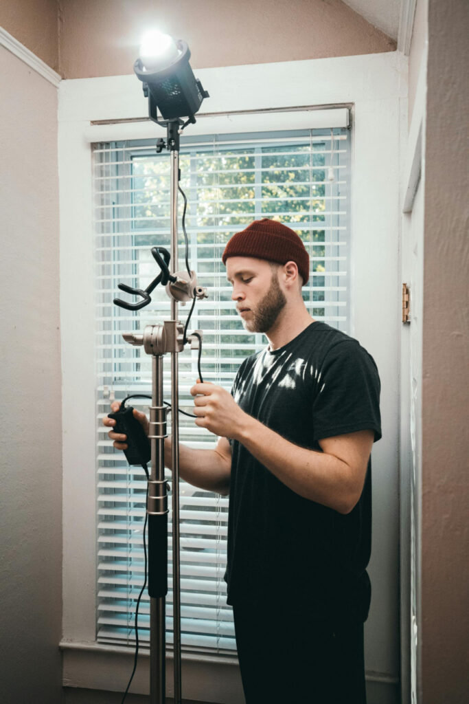 In this image, a man is seen meticulously setting up a professional lighting rig in a well-lit room. He is adjusting the height and angle of the light source, ensuring it is positioned perfectly to enhance the quality of the shoot. The bright light illuminates the space, demonstrating the importance of proper lighting setup in creating high-quality content. This image serves as an excellent visual for the "SETTING UP YOUR LIGHTING" section of the article "THE ULTIMATE GUIDE TO LIGHTING: BOOST YOUR ONLYFANS INCOME NOW!" It highlights the critical steps involved in preparing your lighting equipment to achieve the best results.