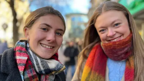 Ellie Whelan (left) and Megan Grimley are standing next to each other, both wearing coats and scarves and smiling at the camera, with a sunny, busy high street in the background.