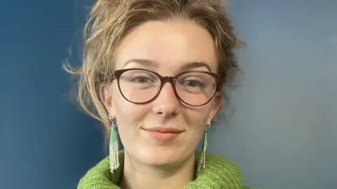 Sarah Peart Sarah Peart is wearing dark-rimmed glasses and a green, knitted roll-neck jumper, and dangly earrings. She has blonde hair which is tied up and is stood against a blue wall.