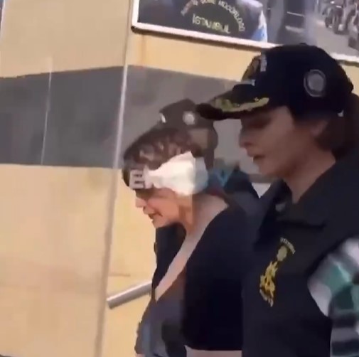 A woman with a bandage on her head is escorted by a police officer.