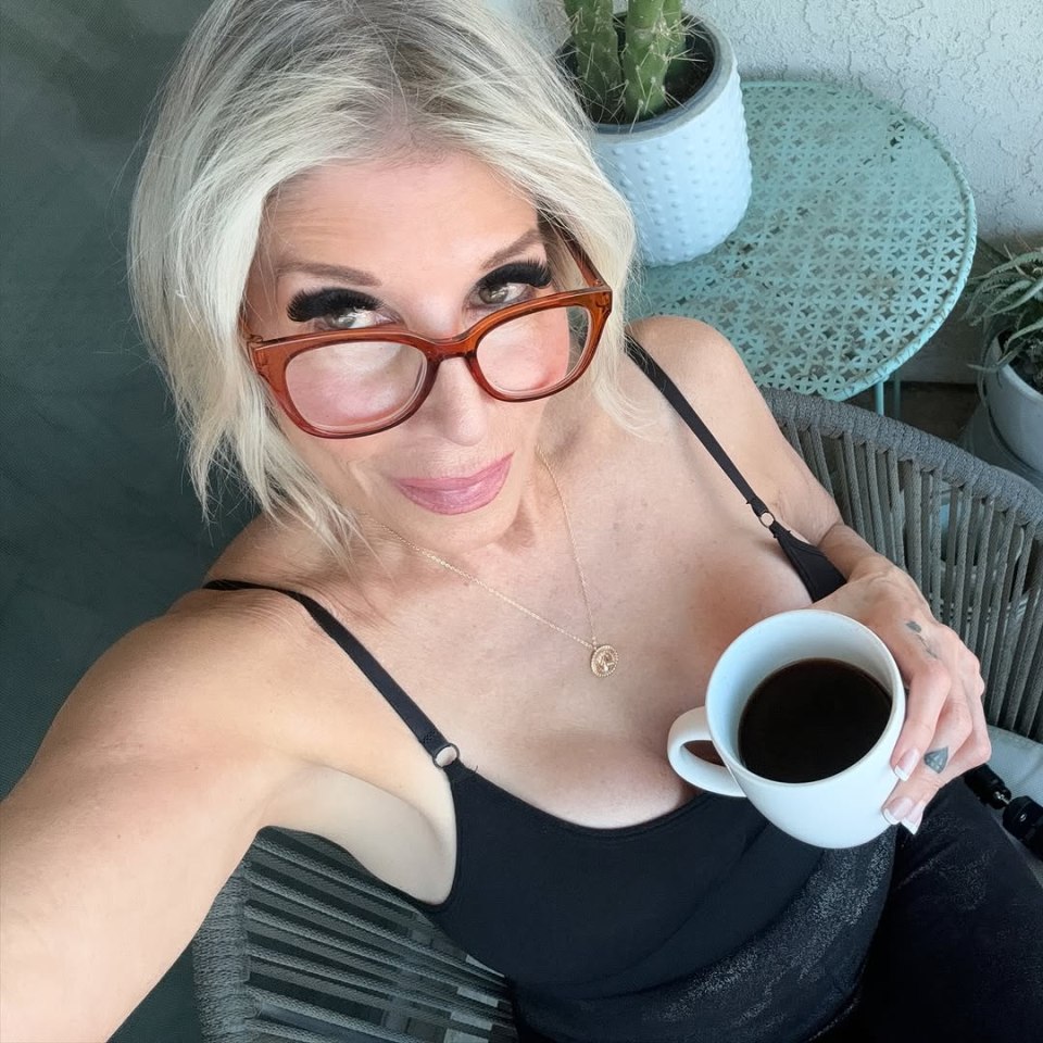 A woman wearing glasses and a black top sits outdoors holding a cup of coffee.