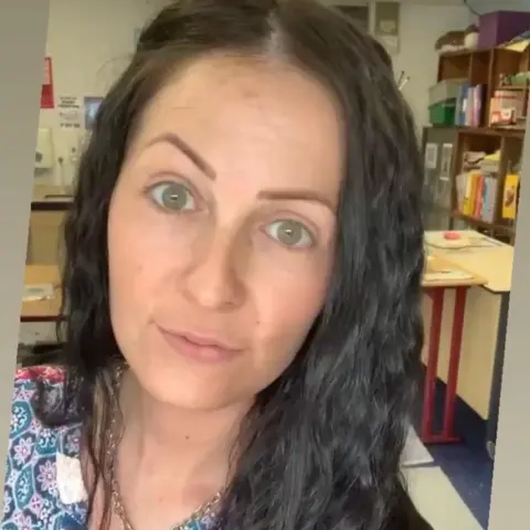 Kirsty Buchan looks into the camera - she has long dark wavy hair and has a blue and purple patterned top. She appears to be in a school - desks, shelves and books can be seen in the background.