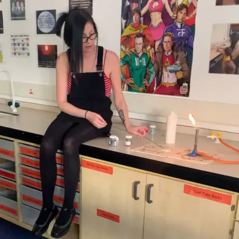 Kirsty Buchan, wearing a black pinafore, black tights and a red striped camisole, sits on a school worktop. To her side is a number of pieces of scientific equipment including a bunsen burner.